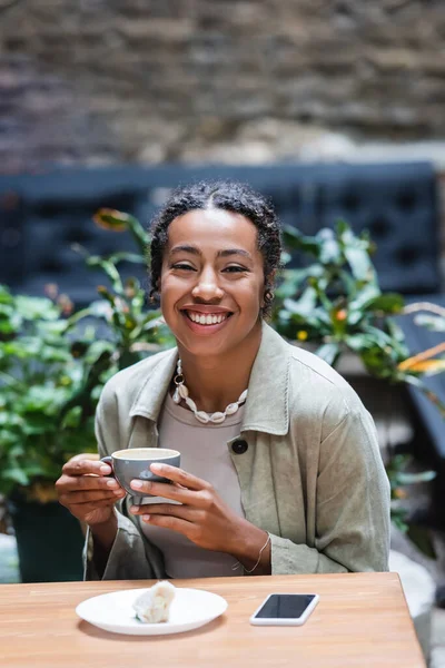 Cheerful african american woman holding cup of coffee near dessert and smartphone in outdoor cafe — Fotografia de Stock