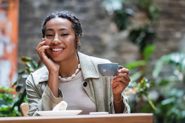 Cheerful african american woman holding cup of coffee near blurred dessert on terrace of cafe — Fotografia de Stock