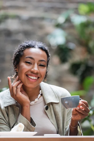 Positive african american woman looking at camera while holding cup of coffee on terrace of cafe — Stock Photo