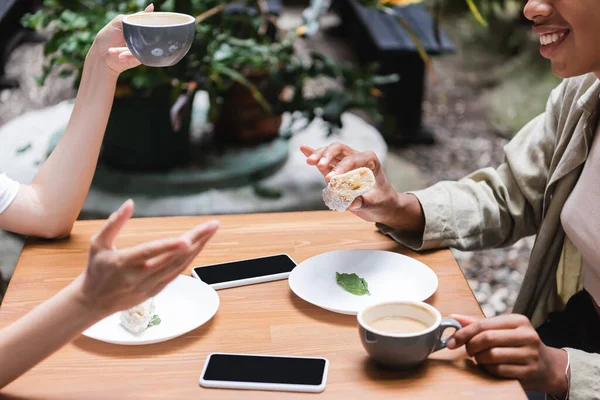 Cropped view of interracial girlfriends spending time near coffee and devices in outdoors cafe — Photo de stock