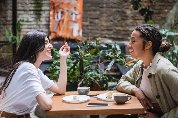 Side view of cheerful multiethnic girlfriends spending time near cellphones and coffee in outdoor cafe — Stock Photo