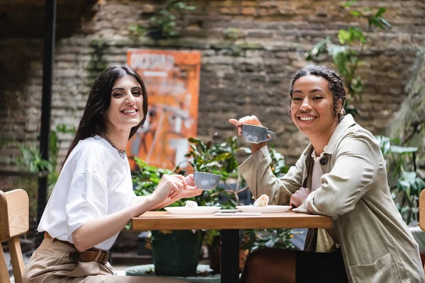 Усміхнені багатоетнічні подружки з кавою дивиться на камеру на терасі кафе — Stock Photo