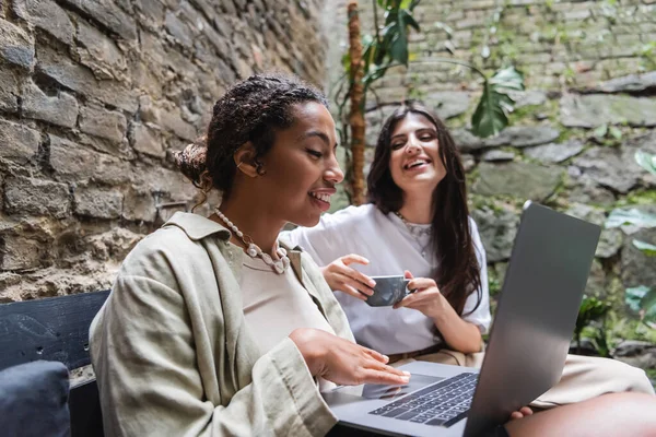 Positive african american woman using laptop near blurred girlfriend with coffee in outdoor cafe — Photo de stock