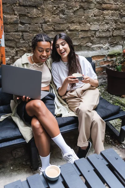 Cheerful interracial girlfriends with coffee using laptop in outdoor coffee — Stock Photo