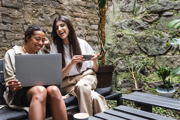 Positive woman holding cup of coffee near african american friend with laptop on terrace of cafe — Photo de stock