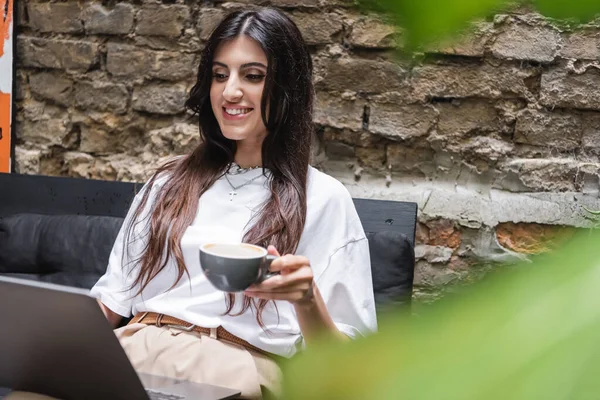 Smiling brunette woman holding cup of coffee and using laptop on terrace of cafe — Stock Photo