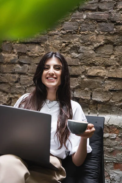 Cheerful brunette freelancer holding cup and laptop while looking at camera on terrace of cafe — Stock Photo