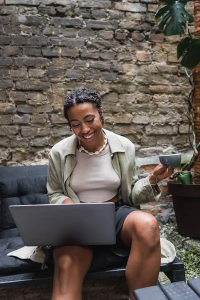 Positive african american woman holding cup and using laptop on terrace of cafe — Stock Photo