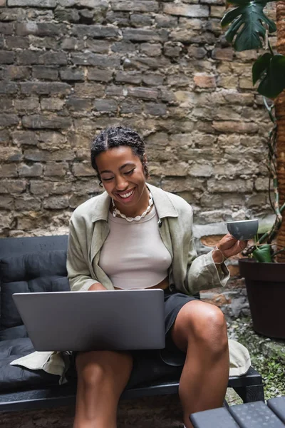 Positive african american woman holding cup and using laptop on terrace of cafe — Stockfoto