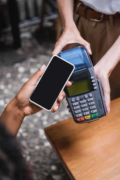 Cropped view of african american client paying with cellphone near waitress on terrace of cafe — Stock Photo