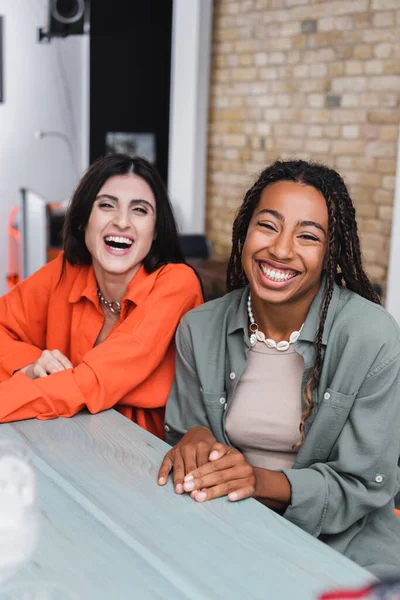 Cheerful interracial girlfriends looking at camera in cafe — Stock Photo
