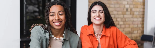 Cheerful interracial girlfriends looking at camera in cafe, banner — Stock Photo