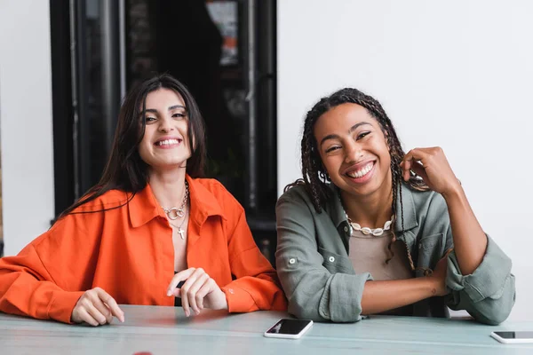 Smiling african american woman looking at camera near friend and smartphones in cafe — Photo de stock