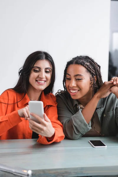 Cheerful african american woman looking at cellphone near friend in cafe - foto de stock