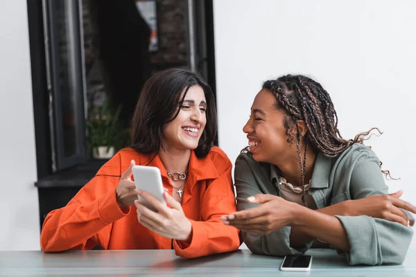 Laughing interracial girlfriends using mobile phone in cafe — Fotografia de Stock