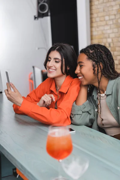 Multiethnic girlfriends using smartphone near cocktail in cafe - foto de stock