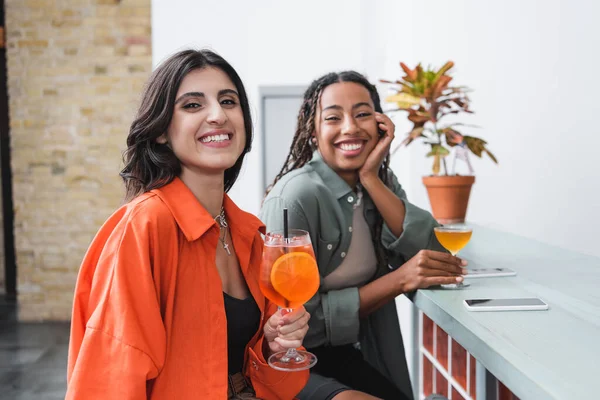 Smiling woman holding cocktail near blurred african american friend and smartphones in cafe - foto de stock