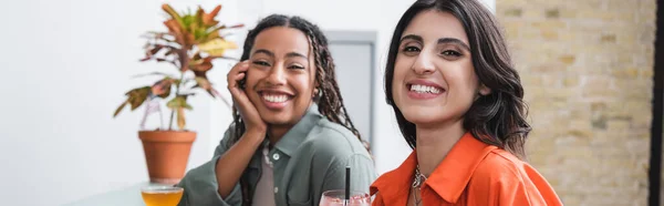 Cheerful woman looking at camera near african american friend and cocktails in cafe, banner — Stock Photo