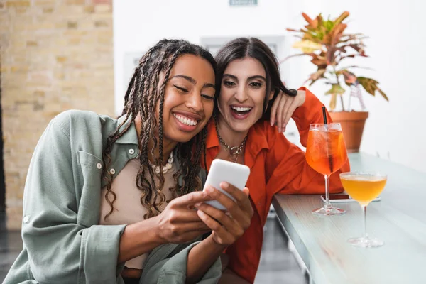 Positive multiethnic friends using smartphone near cocktails in cafe — Stock Photo