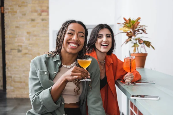 Позитивні міжрасові подружки тримають коктейлі біля смартфонів у кафе — стокове фото