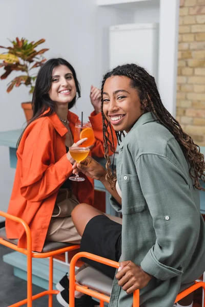 Cheerful african american woman holding cocktail and looking at camera near blurred friend in cafe - foto de stock