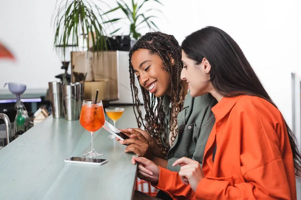 Cheerful multiethnic girlfriends using smartphone near cocktails in cafe — Stockfoto