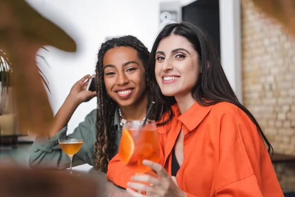 Smiling multiethnic girlfriends with smartphone looking at camera near cocktail in cafe — Stock Photo