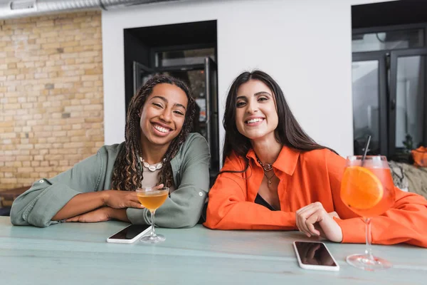 Happy multiethnic girlfriends looking at camera near cocktails and smartphones in cafe — Photo de stock