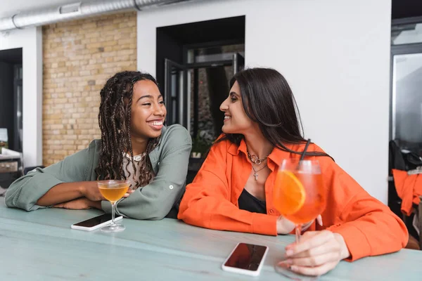 Мультиэтнические девушки разговаривают возле смартфонов и коктейлей в кафе — стоковое фото