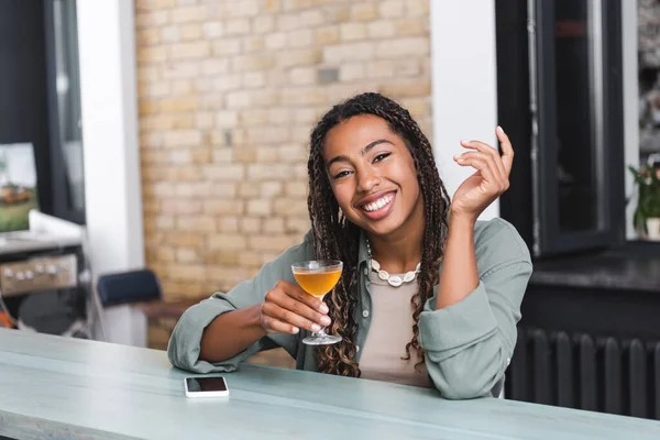 Positive african american woman holding cocktail and looking at camera near smartphone in cafe — Stock Photo