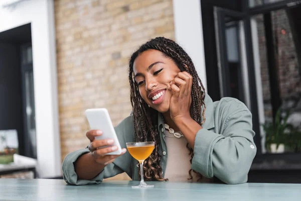 Positive african american woman using mobile phone near cocktail in cafe — Stock Photo