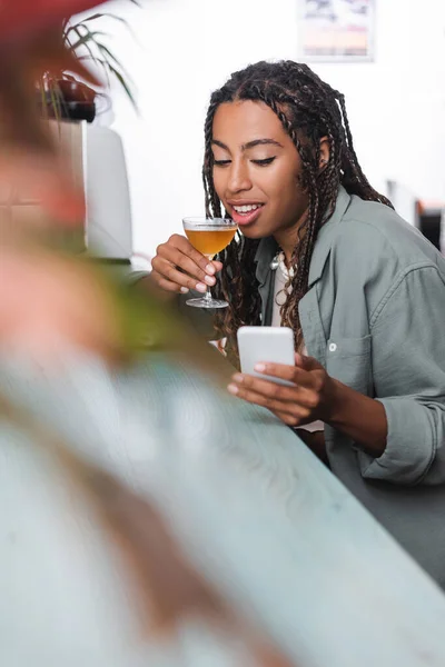 African american woman holding glass of cocktail and using blurred smartphone in cafe - foto de stock