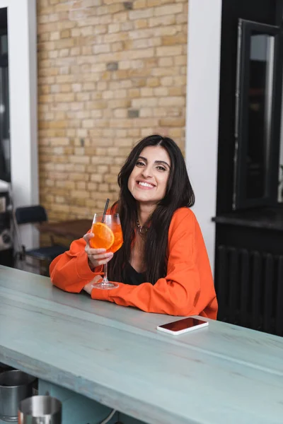 Улыбающаяся женщина держит коктейль возле смартфона в кафе — стоковое фото