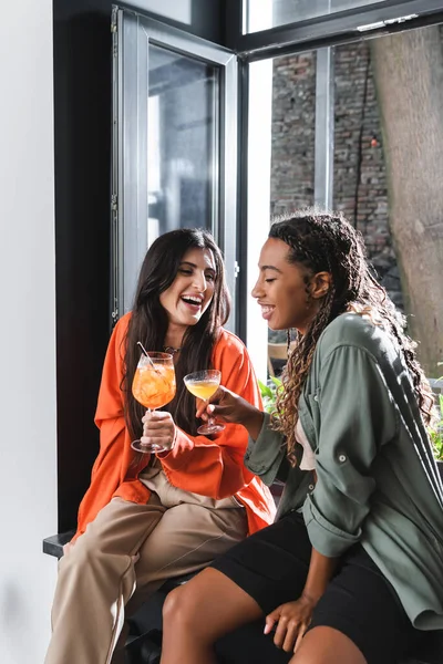 Positive interracial girlfriends holding cocktails on windowsill in cafe — Stockfoto