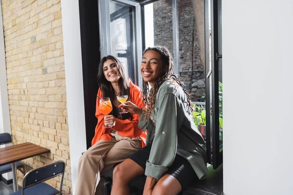 Улыбающиеся многонациональные девушки, держащие коктейли и смотрящие в камеру в кафе — стоковое фото