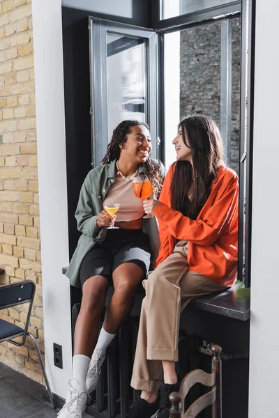 Positive multiethnic friends holding cocktails while sitting on windowsill in cafe — Stockfoto