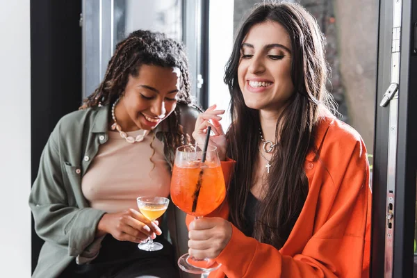 Smiling woman holding cocktail near blurred african american girlfriend and window in cafe — Stockfoto