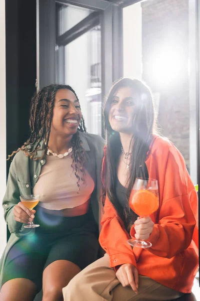 Cheerful interracial girlfriends holding cocktails near window in cafe — Fotografia de Stock