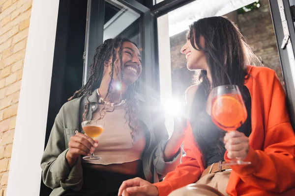 Low angle view of cheerful multiethnic girlfriends with cocktails talking near window in cafe - foto de stock