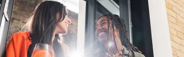 Low angle view of smiling multiethnic girlfriends talking near cocktail in cafe, banner — Foto stock