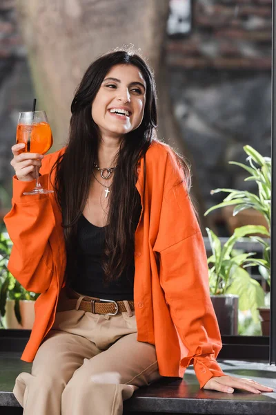 Positive woman holding cocktail and looking away on windowsill in cafe — Stockfoto