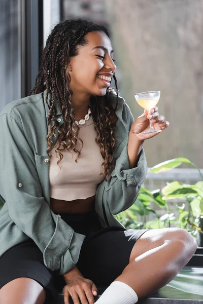 Smiling african american woman holding cocktail while sitting on windowsill in cafe — Foto stock