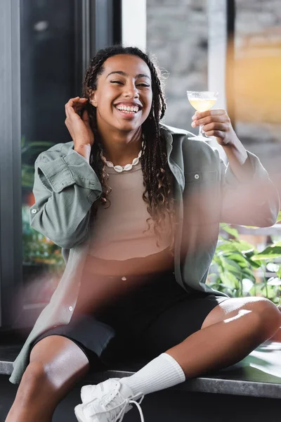 Happy african american woman holding cocktail while sitting near window in cafe — Foto stock