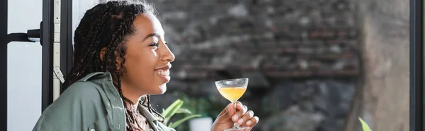 Side view of positive african american woman holding cocktail near window in cafe, banner — Photo de stock