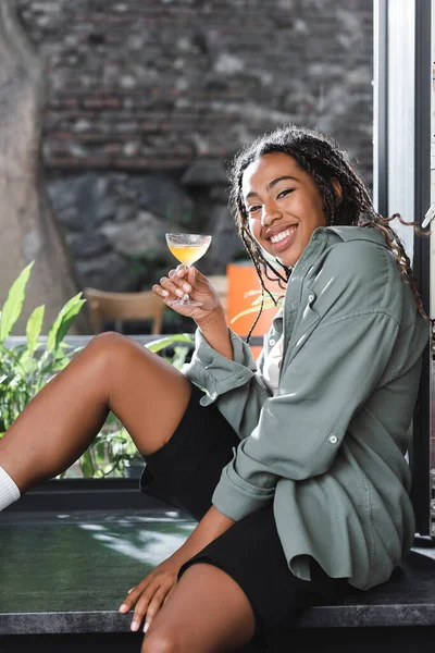 Positive african american woman holding cocktail and looking at camera on windowsill in cafe - foto de stock