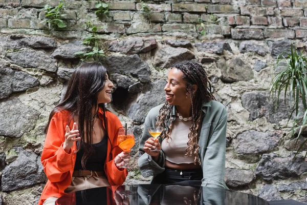 Smiling interracial girlfriends with cocktails talking on terrace of cafe — Photo de stock