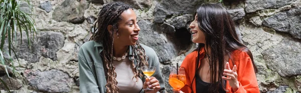 Positive multiethnic girlfriends holding cocktails while talking on terrace of cafe, banner — Foto stock