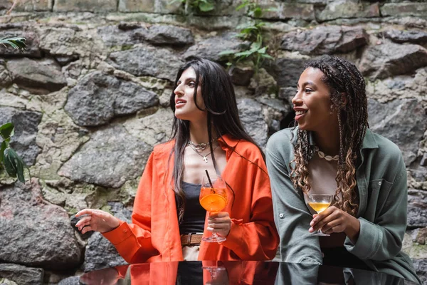 Багатоетнічні подружки тримають коктейлі і дивляться на терасу кафе — стокове фото
