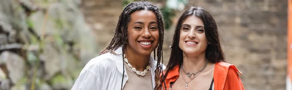 Interracial girlfriends smiling at camera on terrace of cafe, banner - foto de stock