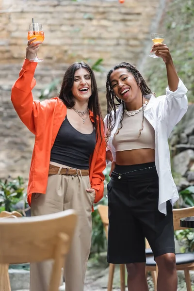 Excited multiethnic women holding cocktails and looking at camera on terrace of cafe — Fotografia de Stock